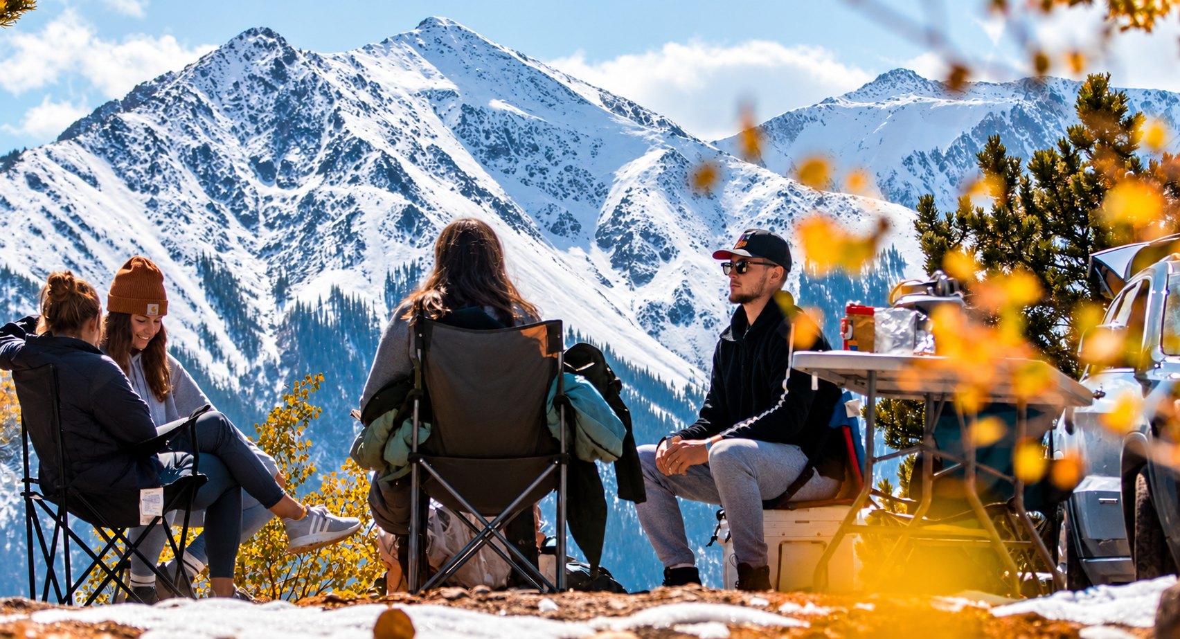 Travellers relaxing at a mountain overlook beneath snowy peaks and autumn trees
